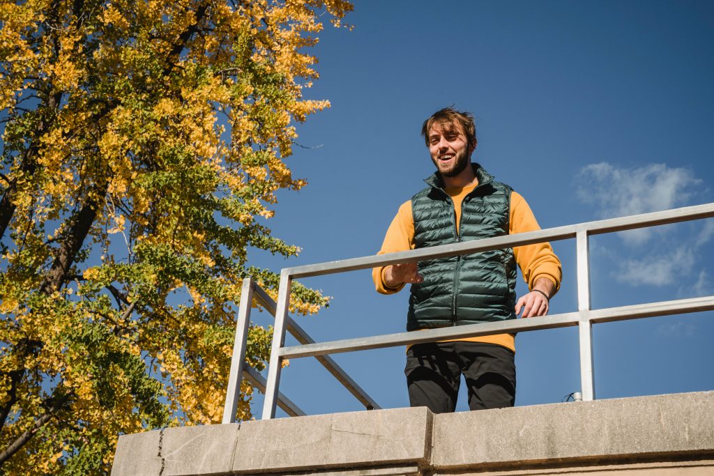 Low angle of cheerful bearded male holding hand on fence while standing on height and looking down